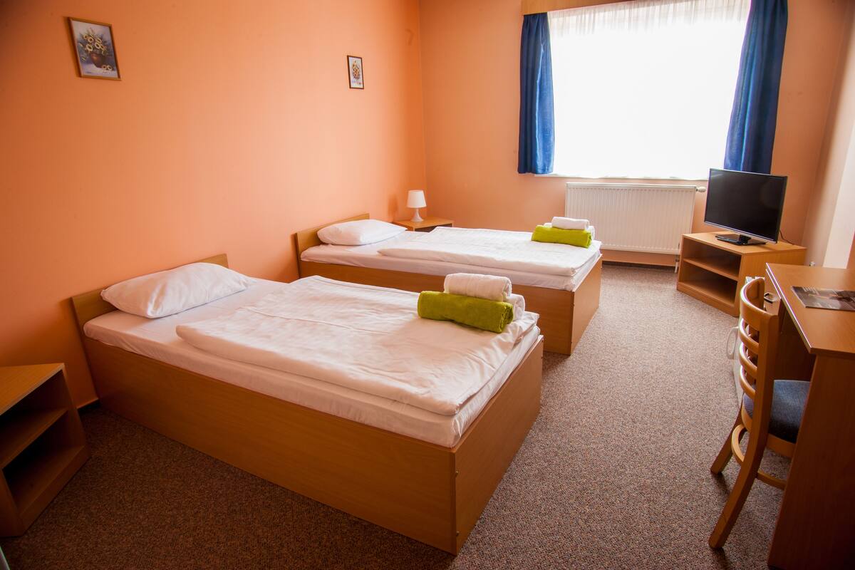Quarto de hotel com duas camas de solteiro, uma escrivaninha com cadeira, televisão e uma janela com cortinas.