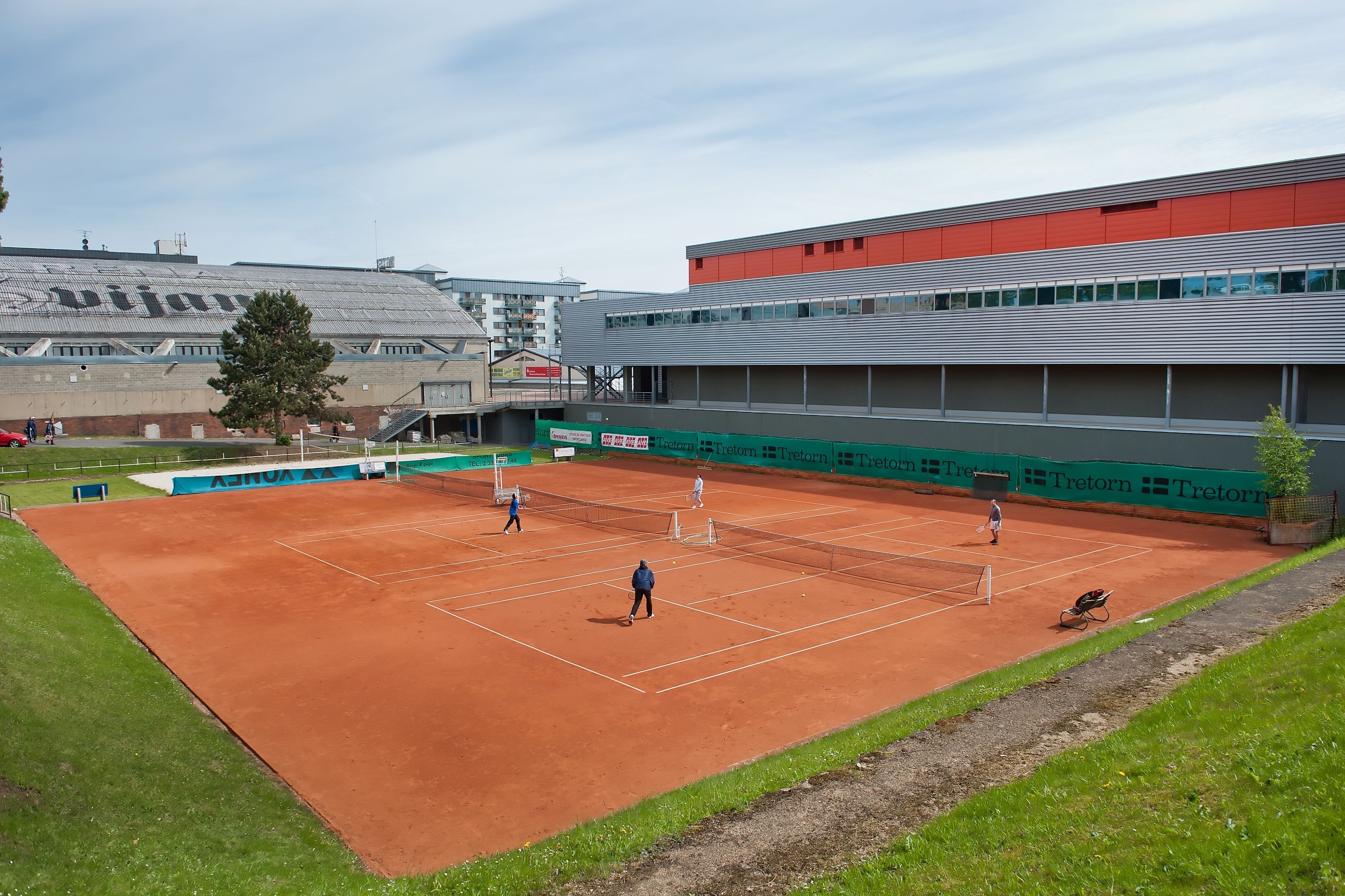 Een tennisbaan met spelers, omringd door een gebouw met een rood dak en reclame.