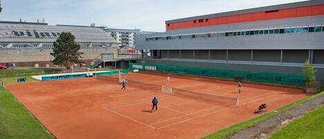 Cancha de tenis con jugadores, rodeada por un edificio con tejado rojo y anuncios.
