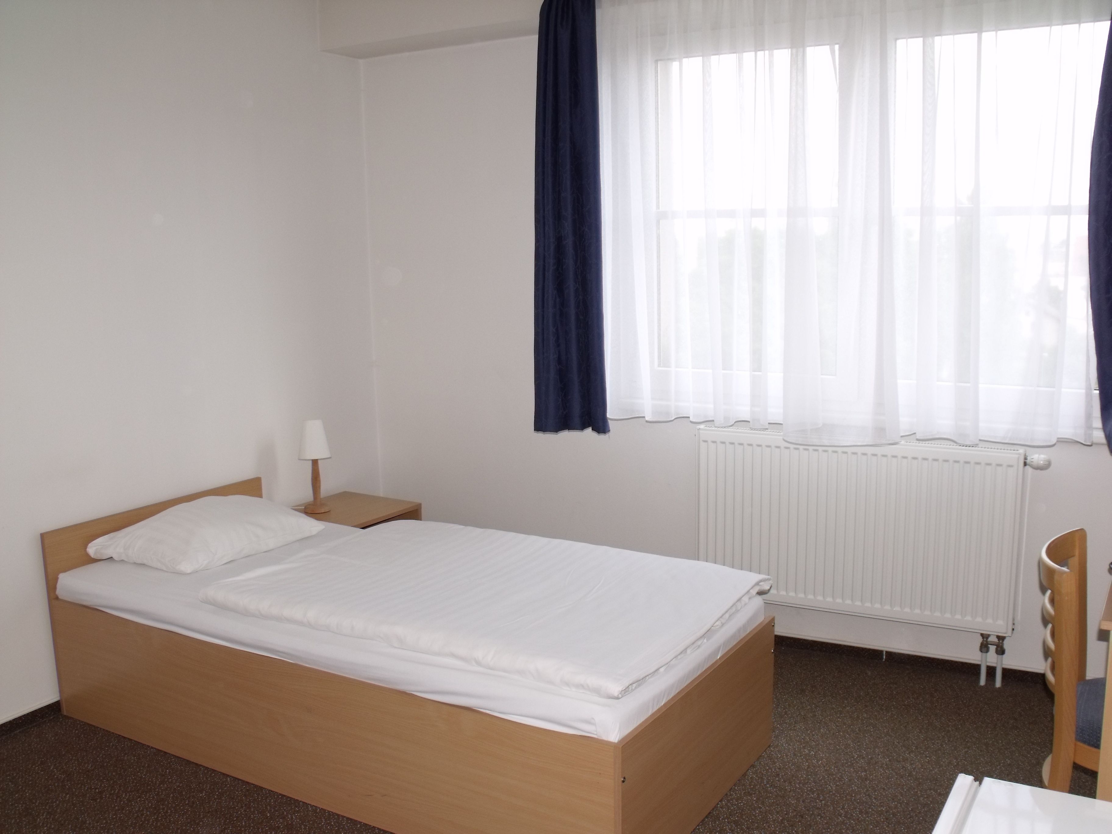 A simple, clean room with a single bed, a nightstand, a chair, and a window with sheer curtains.