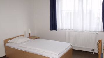 A simple, clean room with a single bed, a nightstand, a chair, and a window with sheer curtains.