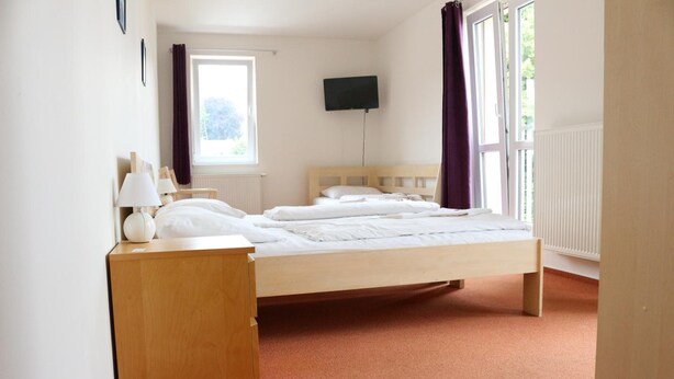 A bedroom with a bed, a bedside table, a lamp, a TV, and a window with curtains. - Hotel Petra (Liberec)