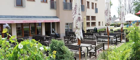 An outdoor seating area with tables and chairs, surrounded by greenery and a building with multiple windows and balconies.