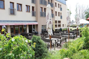 Un área de estar al aire libre con mesas y sillas, rodeada de vegetación y un edificio con múltiples ventanas y balcones.