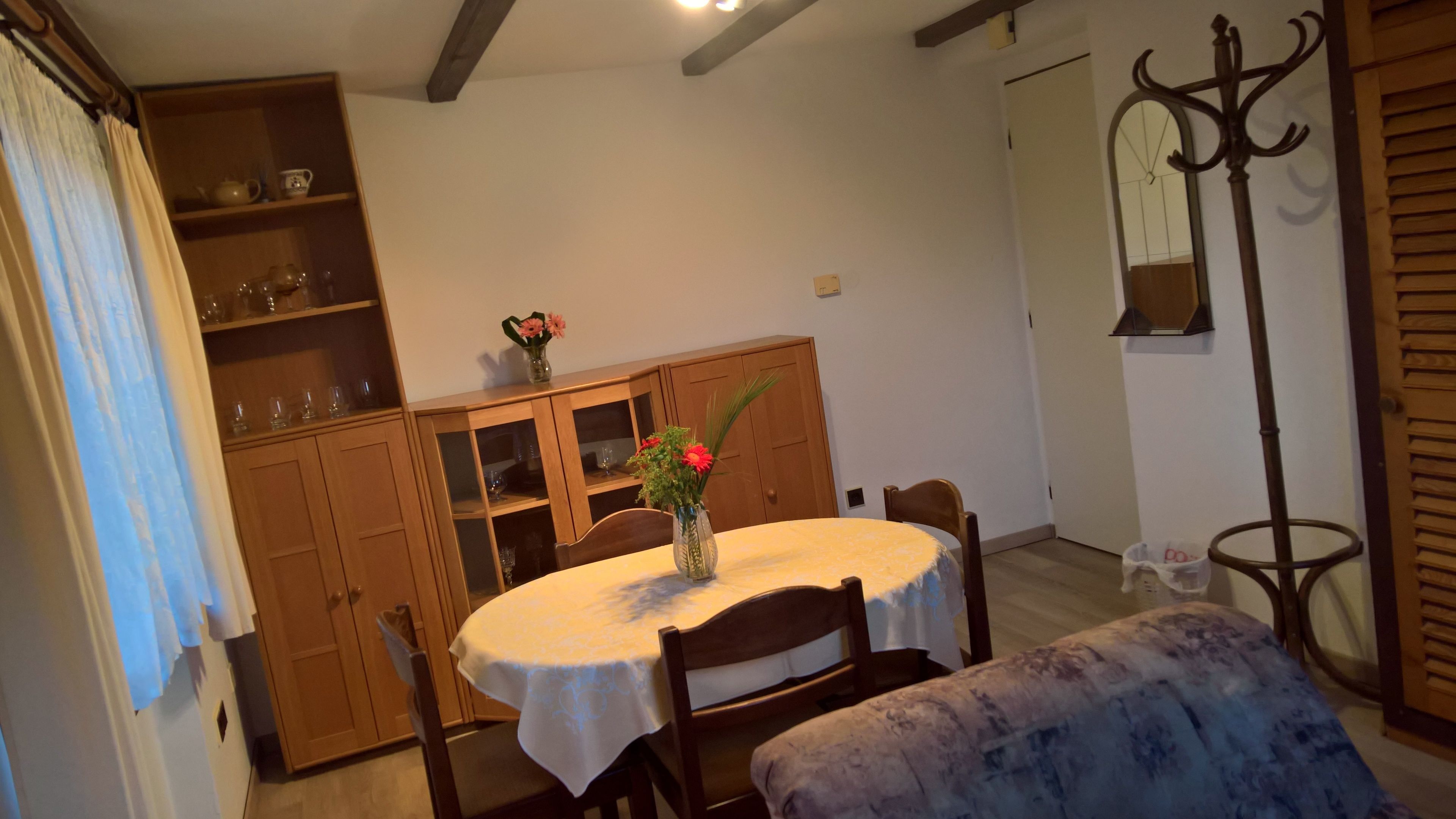 A dining area with a round table, chairs, and a vase with flowers. There is a wooden cabinet with glass doors and a coat rack.
