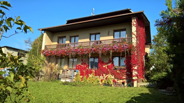 A two-story building with a balcony adorned with red and pink flowers, surrounded by greenery.