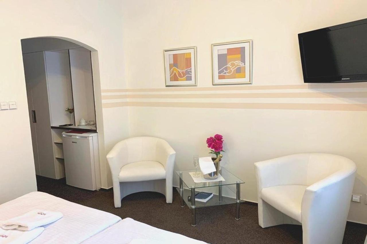 Ein Hotelzimmer mit einem Bett, zwei weißen Sesseln, einem kleinen Glastisch mit Blumenvase, einem Flachbildfernseher und einem Kunstwerk an der Wand.