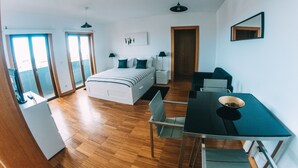 Ein modernes Schlafzimmer mit Bett, Nachttischen, einem Schreibtisch und einem Sessel. Fenster und eine Tür sind zu sehen.