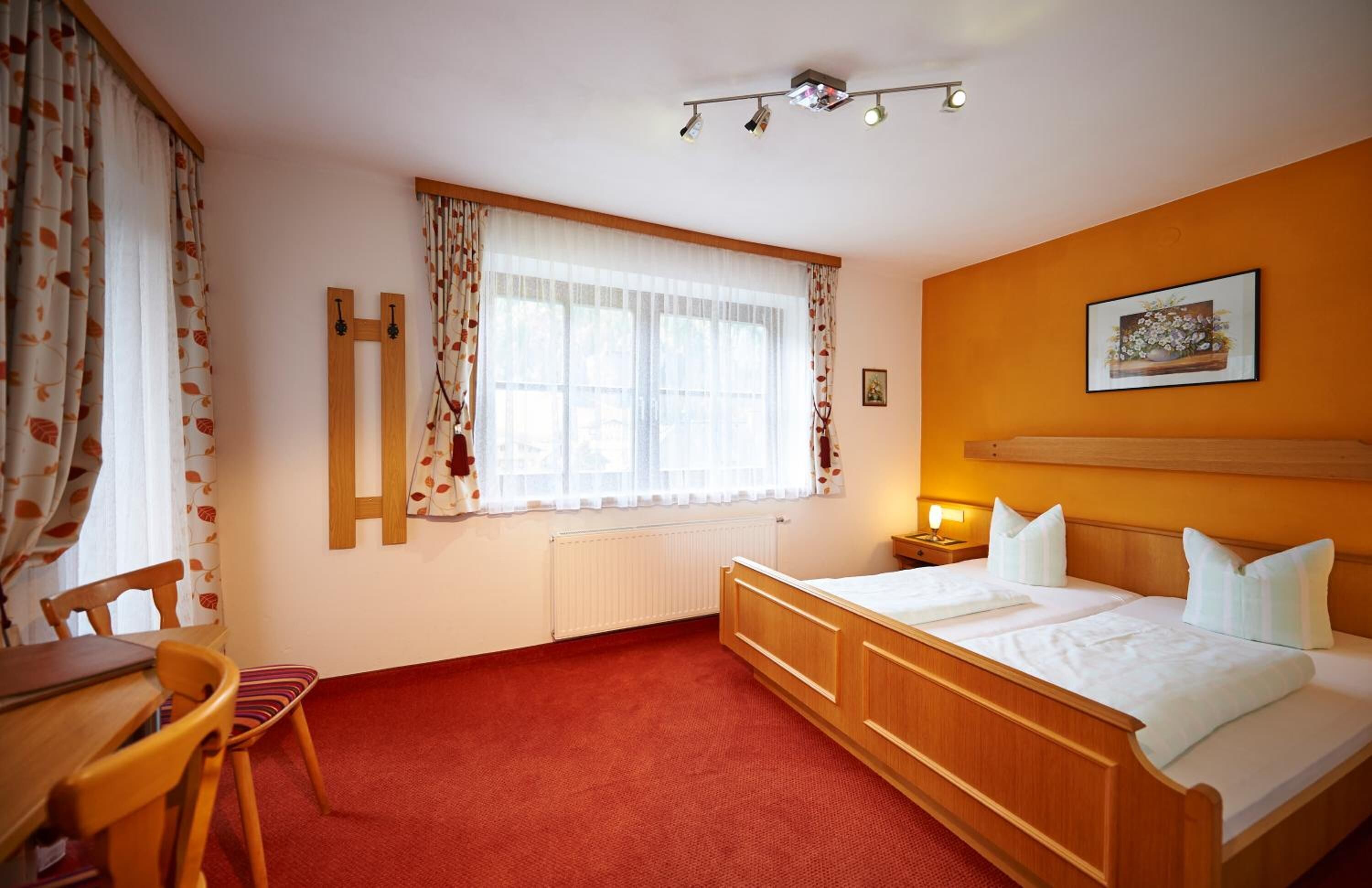 A bedroom with a wooden bed, a desk, and a chair. There is a window with curtains, a radiator, and a picture on the wall.