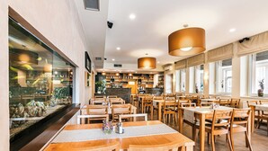 Interior de un restaurante con mesas y sillas de madera, una zona de bar y grandes ventanales.