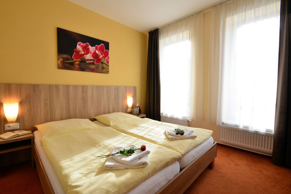 Ein Hotelzimmer mit zwei Betten, einem Holz-Kopfteil, einem Nachttisch mit Lampe, einem Blumenbild und einem Fenster mit Vorhängen.