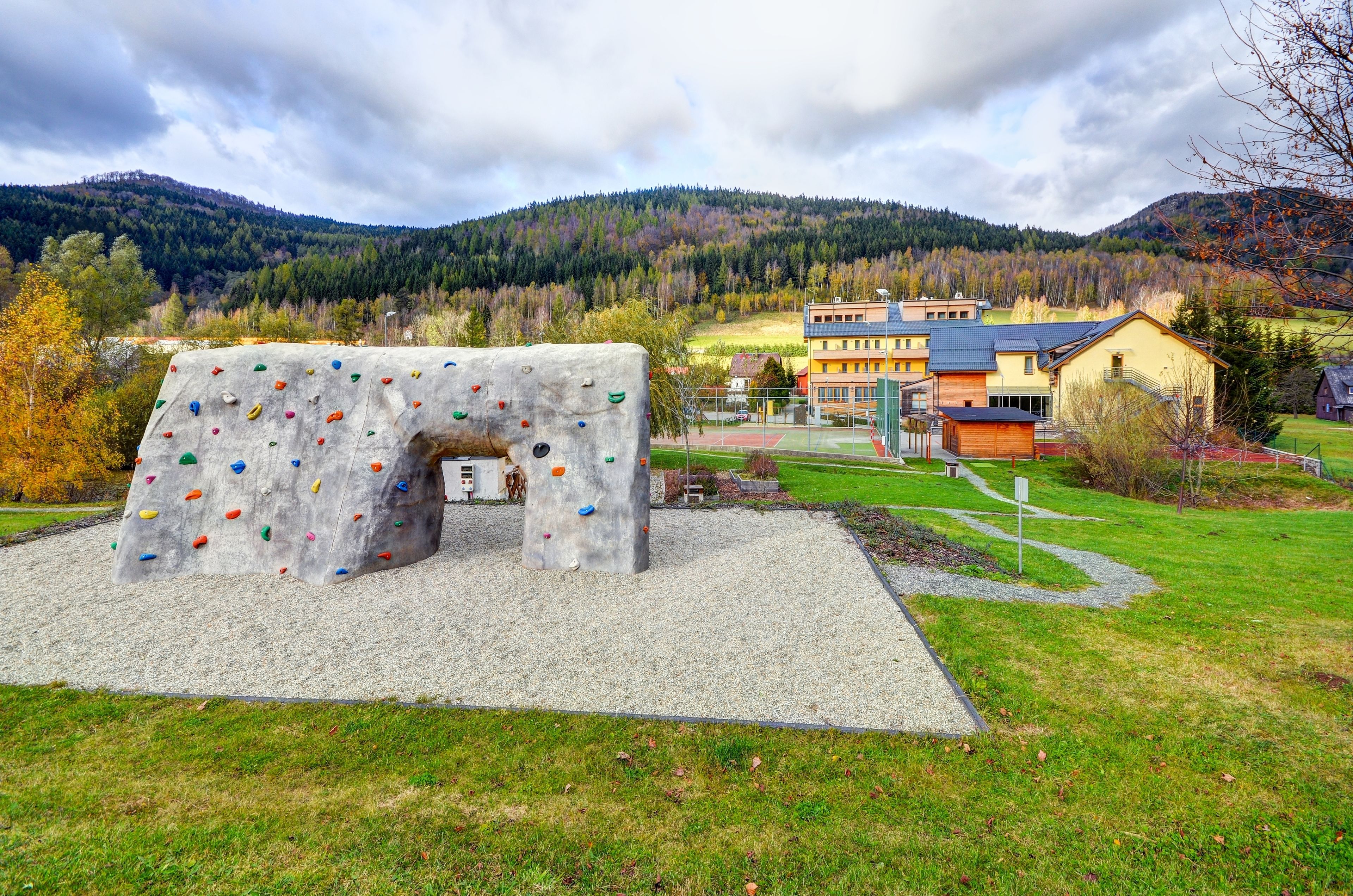A climbing wall with colorful holds in a park setting, surrounded by a building with a red roof and a tennis court.