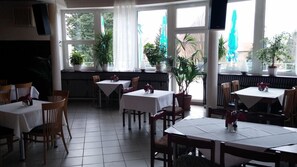 Un restaurante con varias mesas y sillas, amplias ventanas con cortinas y plantas en macetas.