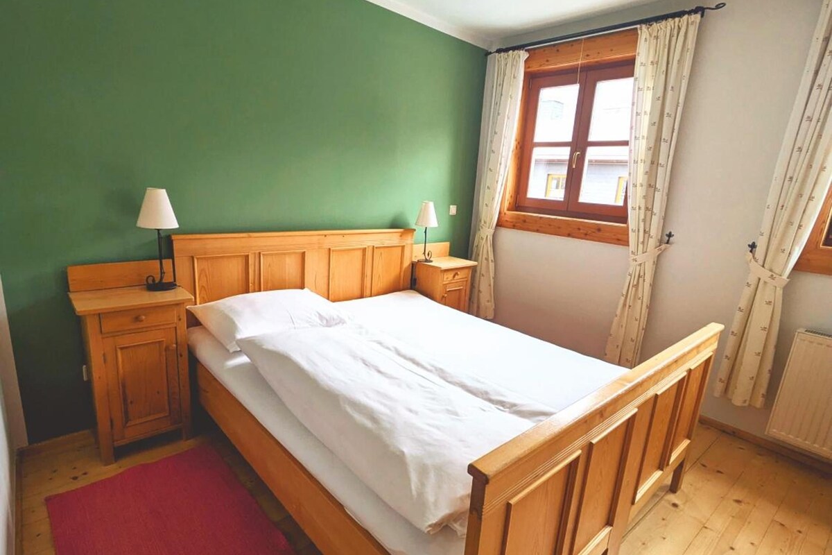 Une chambre avec un lit simple, une tête de lit en bois, un tapis rouge, une table de chevet avec une lampe et une fenêtre avec des rideaux.