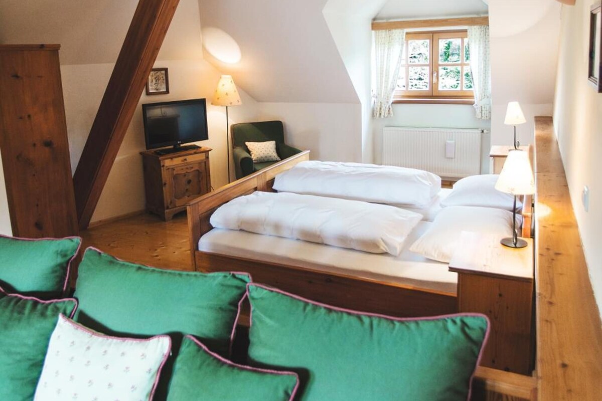 Une chambre avec un plafond en pente, un lit avec une literie blanche, un canapé vert, une table de chevet en bois, une lampe et une fenêtre avec des rideaux.