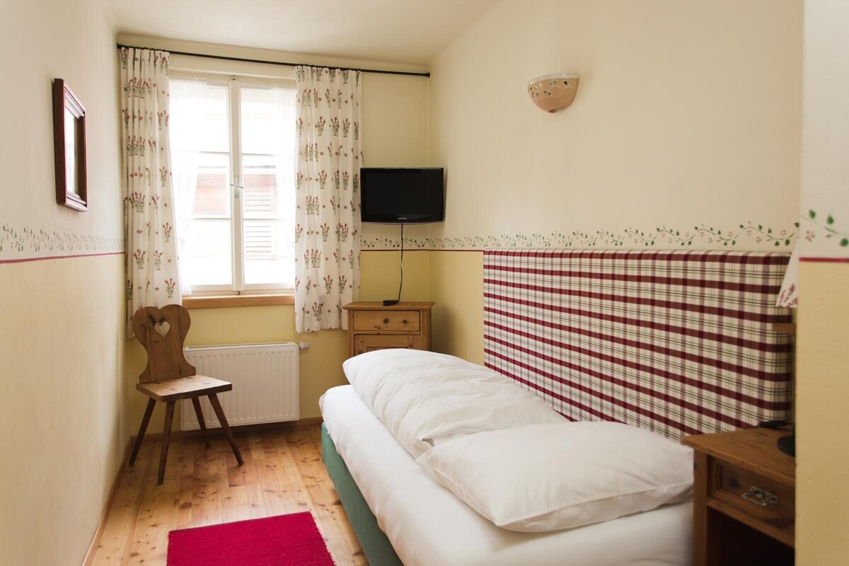 Une chambre à coucher avec un lit, une chaise, une télévision et une fenêtre avec des rideaux.