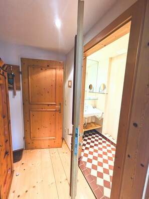 Une porte en bois donne sur une salle de bain dont le sol est carrelé et où se trouve un miroir au-dessus du lavabo.