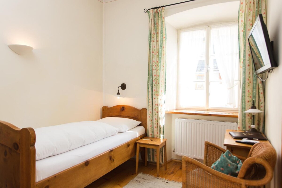 Une petite chambre avec un lit simple, une tête de lit en bois, une table de chevet, une chaise, un bureau avec une lampe, un téléviseur à écran plat fixé au mur et une fenêtre ornée de rideaux à motifs floraux.