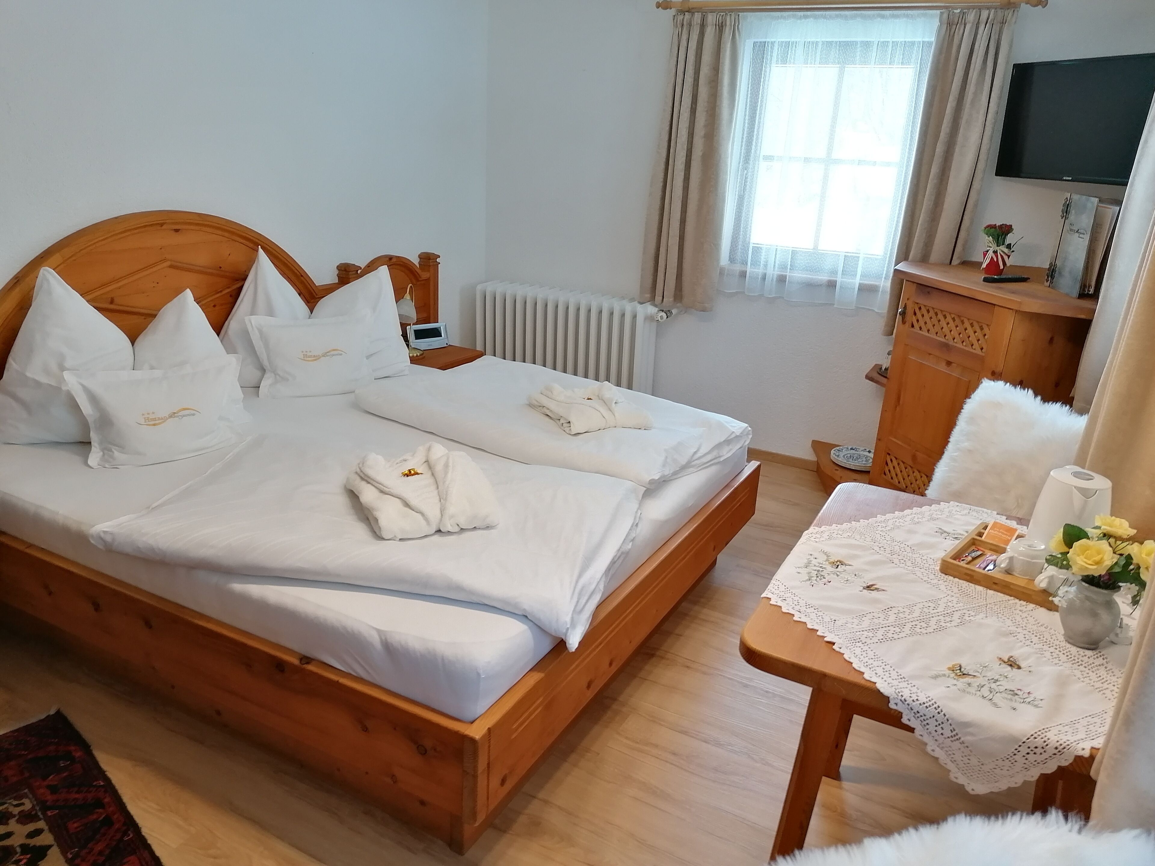 A hotel room with a wooden bed, white bedding, a bedside table with a vase of flowers, and a view of the outside through a window with curtains.