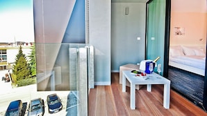 Habitación de hotel con balcón, una cama, una mesita con una botella y vasos, y vistas a coches aparcados y un edificio.