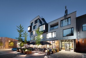 Um edifício moderno com um terraço de restaurante, mesas ao ar livre e entrada visível.