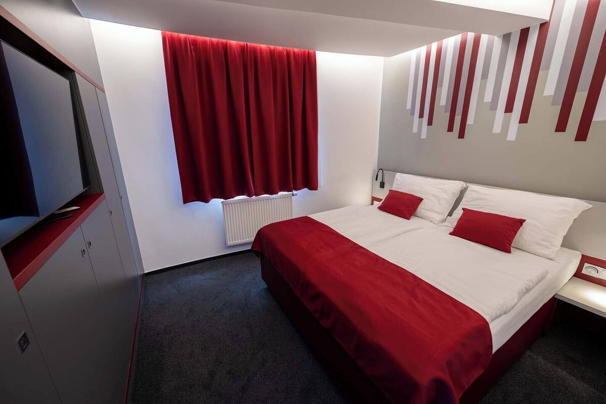Une chambre d’hôtel avec un grand lit, des rideaux rouges, un radiateur blanc et un téléviseur fixé au mur.
