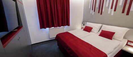 A hotel room with a large bed, red curtains, a white radiator, and a wall-mounted TV.