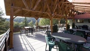 An outdoor patio with wooden beams, tables, and chairs, overlooking a mountainous landscape.