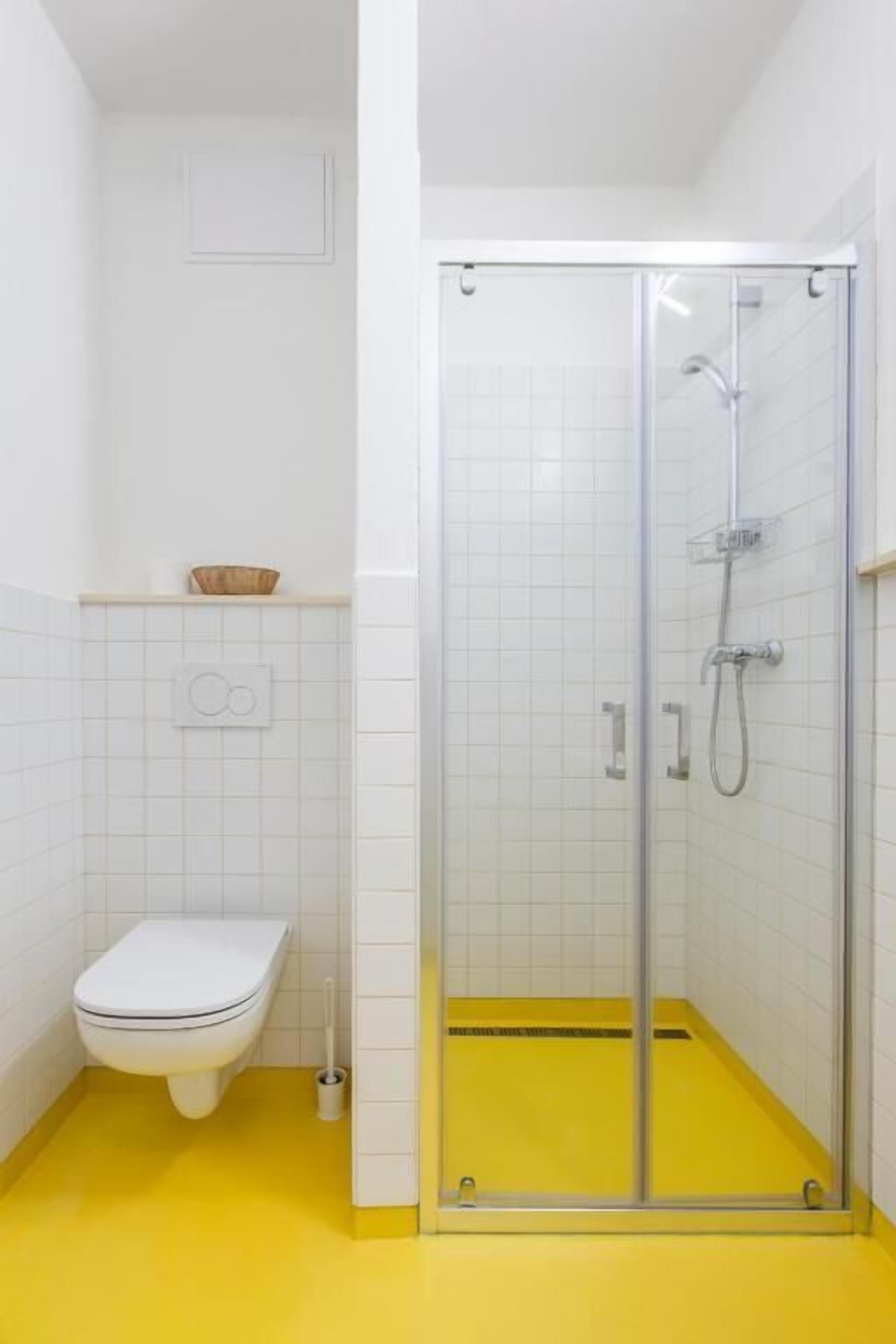 Et badeværelse med et hvidt toilet, en glasvægs brusekabine og gulv i gult.
