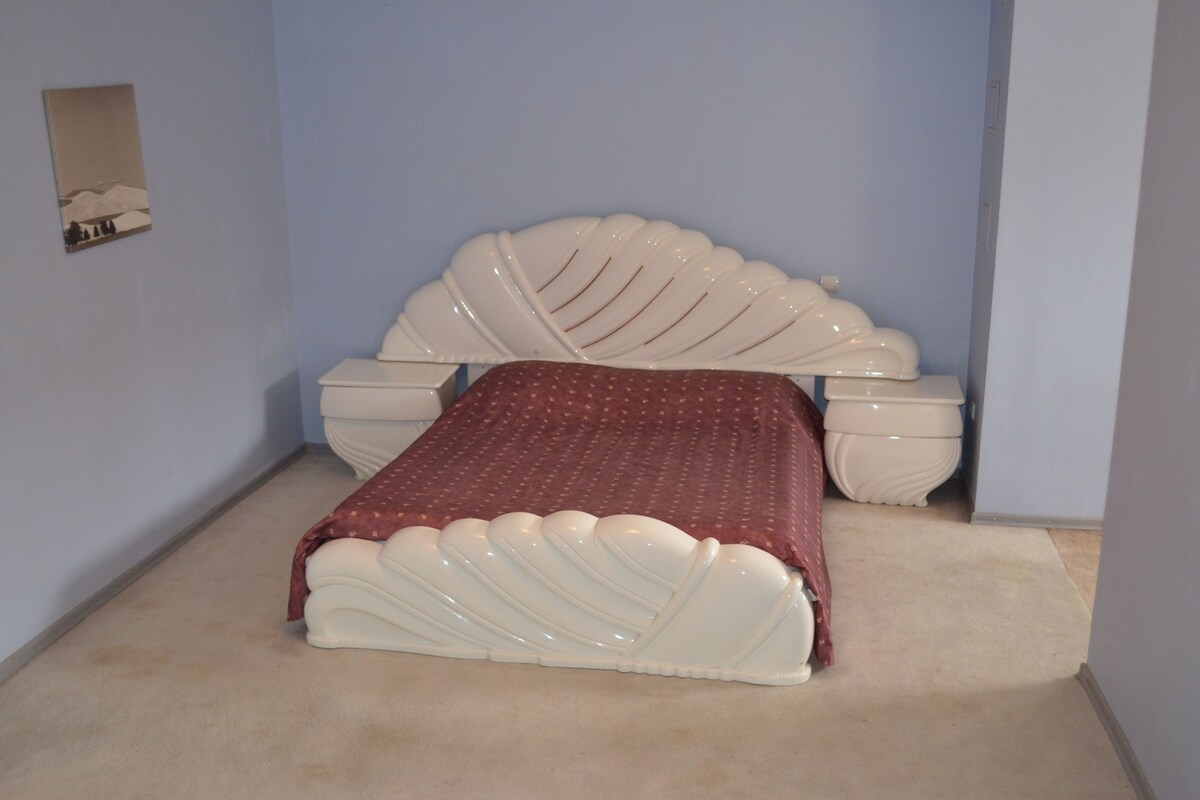 Un dormitorio con un diseño de cama original, una colcha roja y una mesita de noche pequeña.
