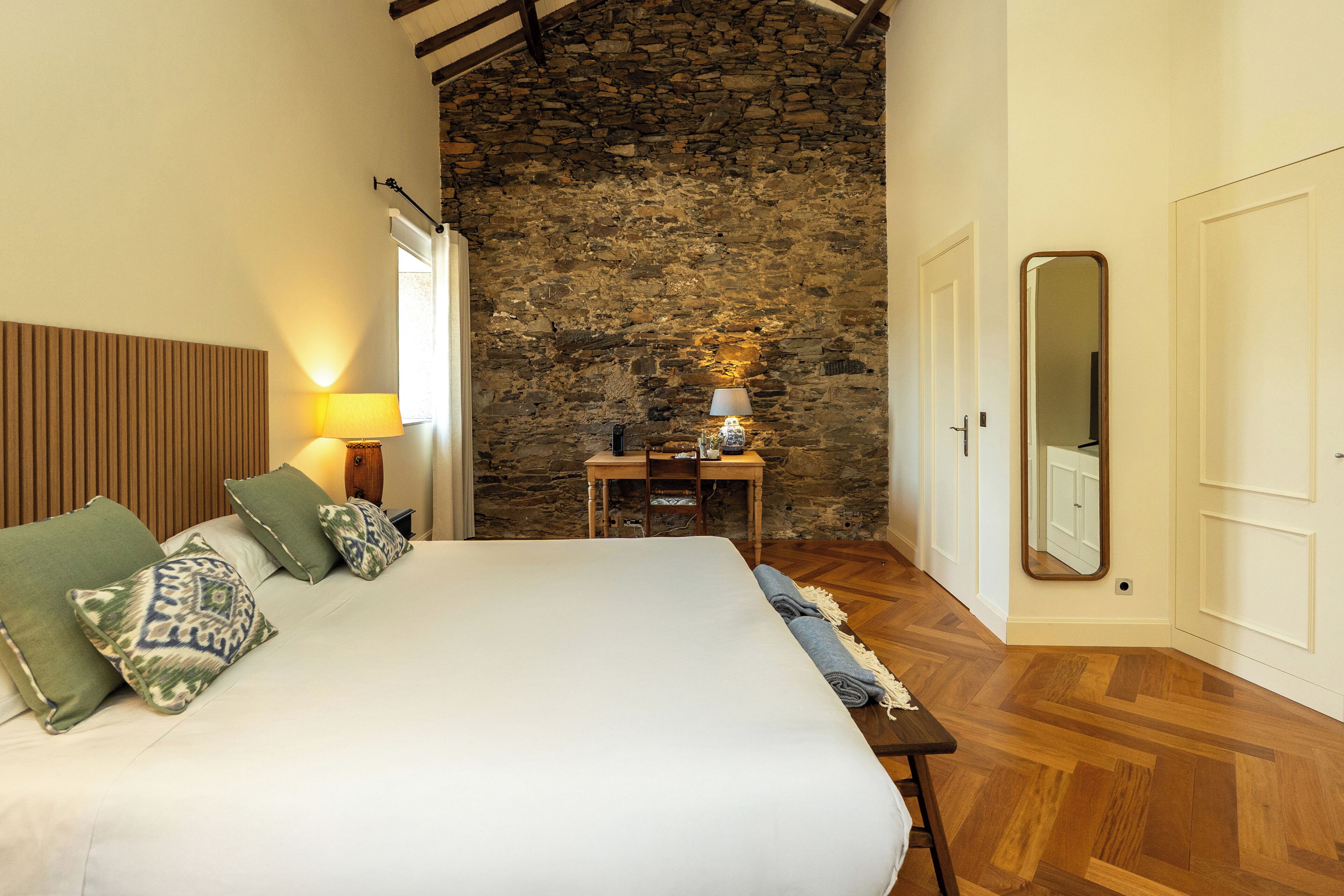 Un dormitorio con una cama grande, cabecera de madera, pared de piedra, una mesita con lámpara y un espejo en la pared.