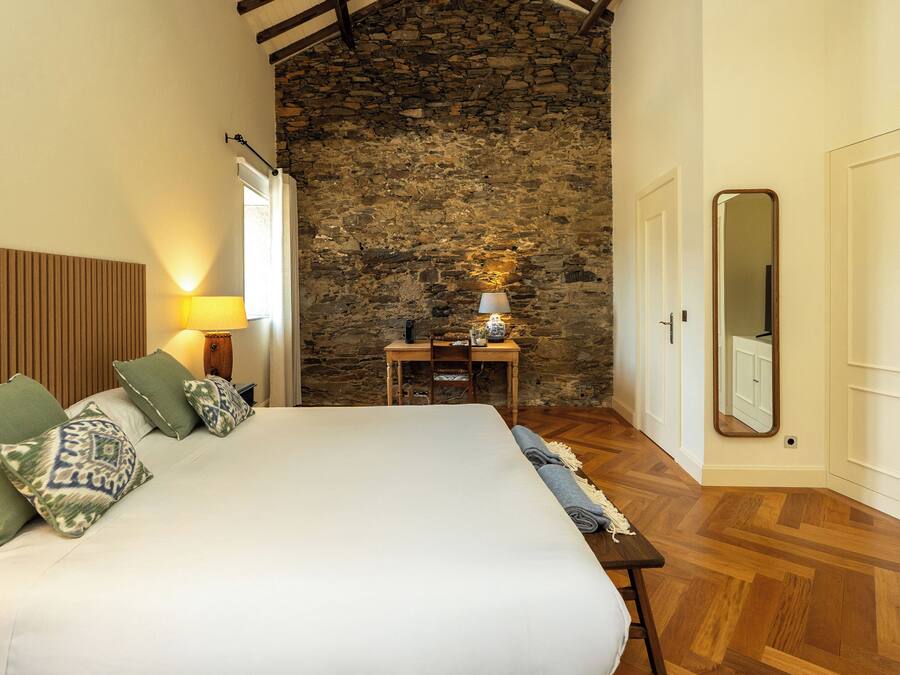 Un dormitorio con una cama grande, cabecera de madera, pared de piedra, una mesita con lámpara y un espejo en la pared.