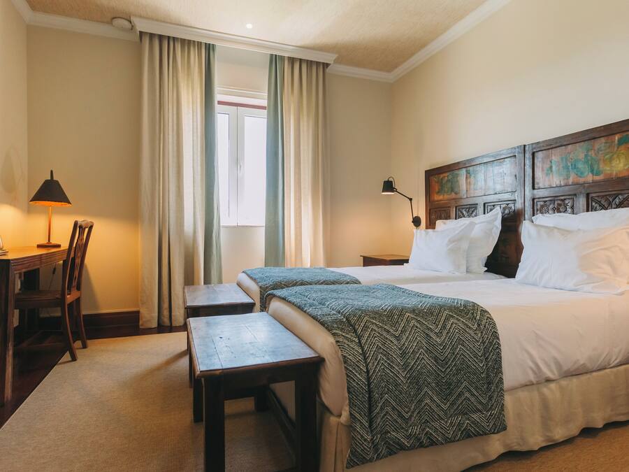 Une chambre d’hôtel avec deux lits, un bureau, une chaise, une lampe et une fenêtre avec des rideaux.