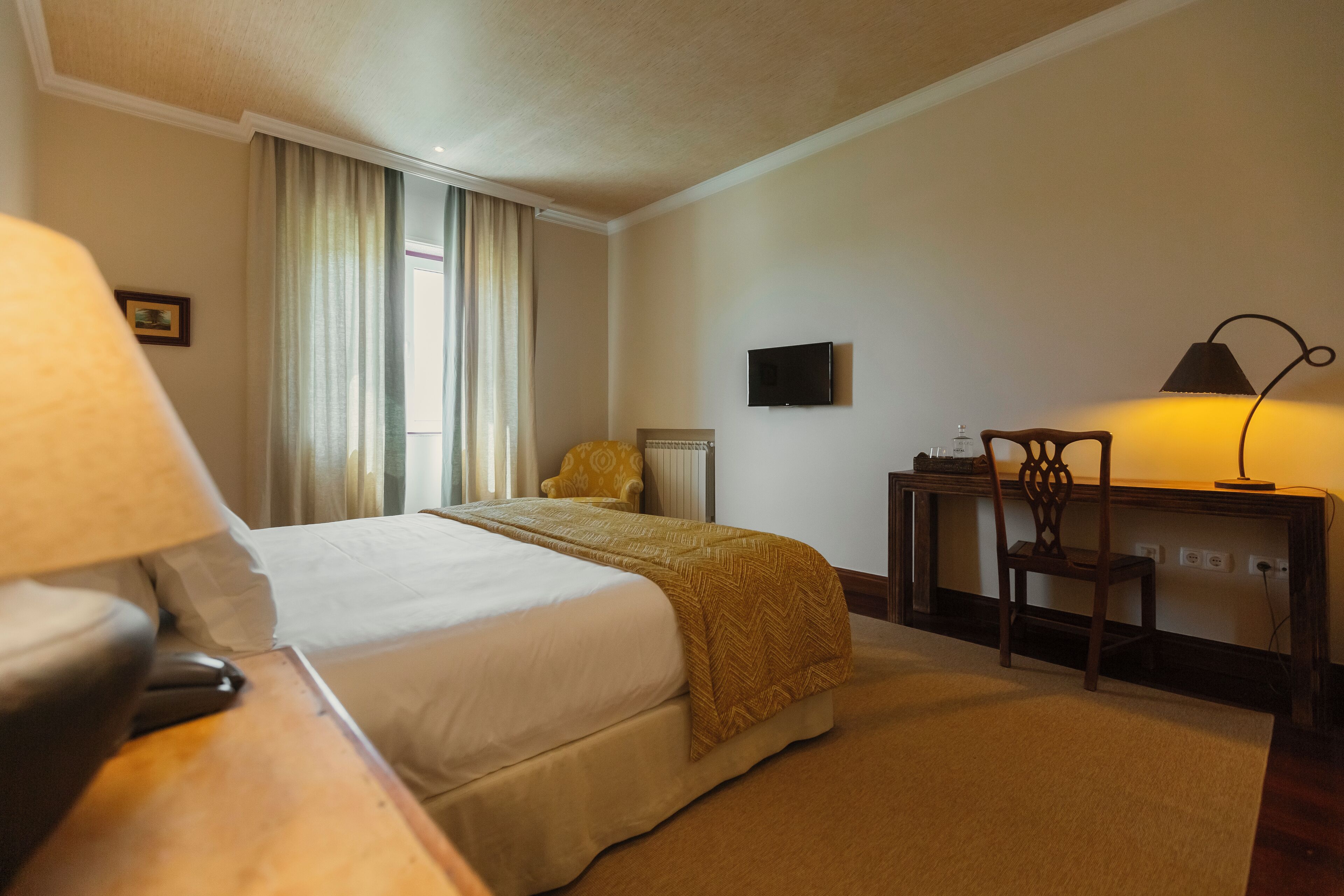 A hotel room with a bed, a desk, a chair, a lamp, and a television on the wall.