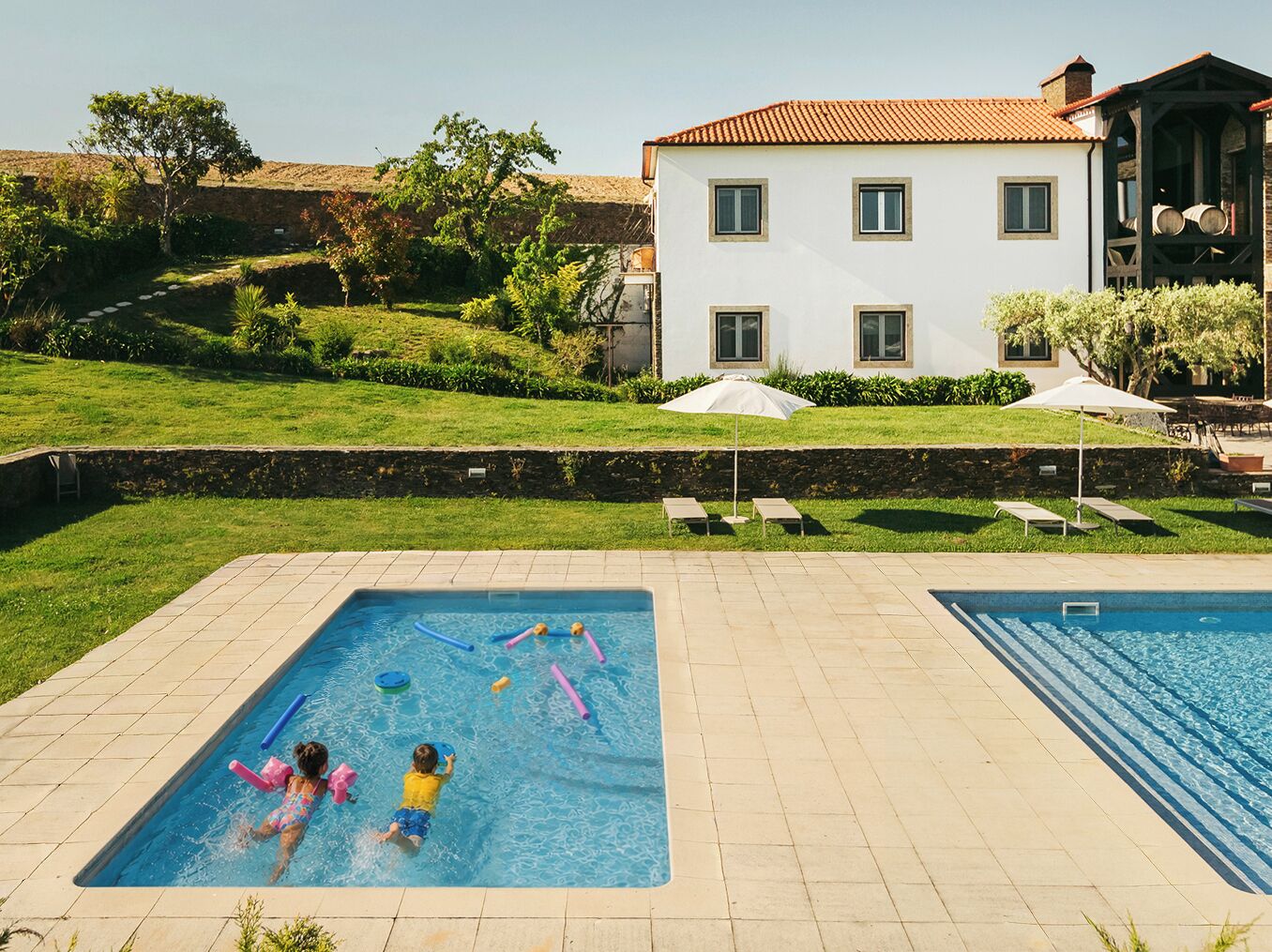 Ein Poolbereich mit zwei Schwimmbecken, einer Rasenfläche und einem Gebäude mit rotem Dach im Hintergrund.
