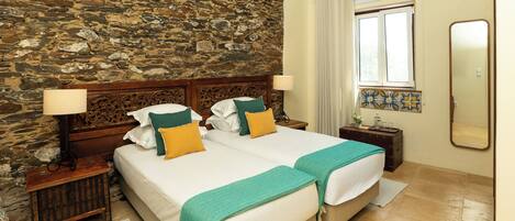 A double bed with two pillows, a nightstand with a lamp, a stone wall, a window with curtains, and a mirror on the wall.