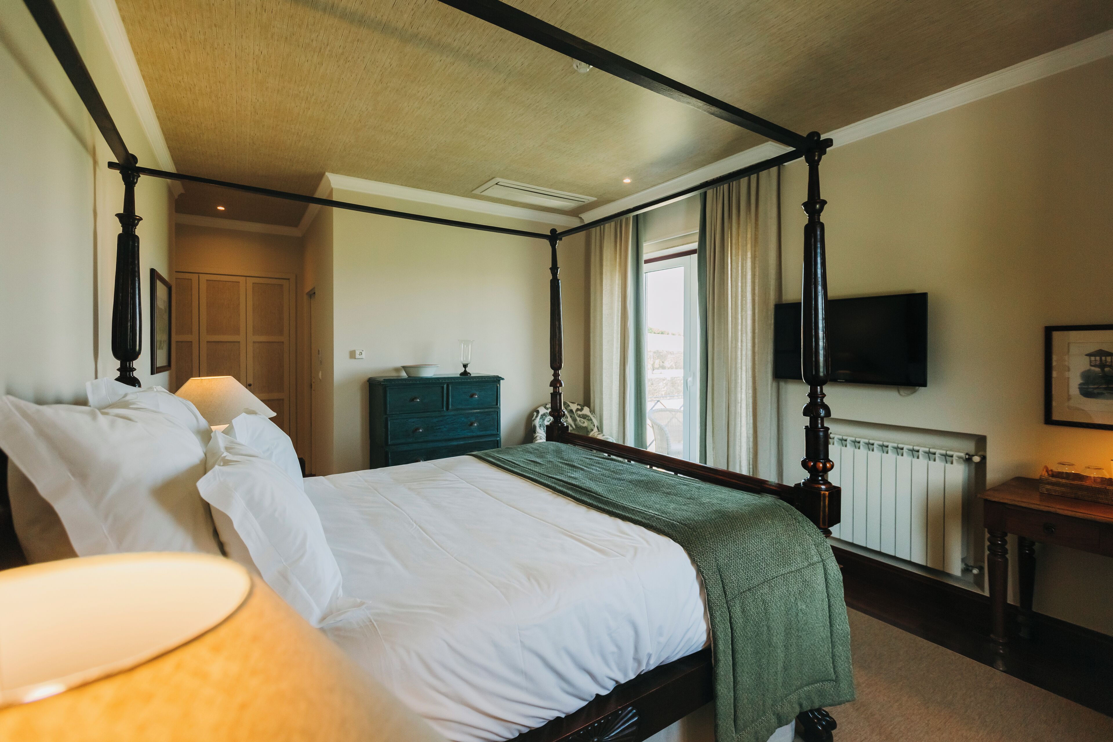 A four-poster bed with white linens, a green throw blanket, and a bedside table with a lamp. A television is mounted on the wall, and a window with curtains is visible.