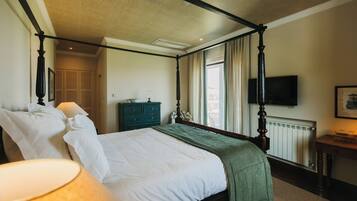 Uma cama de dossel com lençóis brancos, um cobertor verde e uma mesa de cabeceira com abajur. Uma televisão está montada na parede, e é visível uma janela com cortinas.