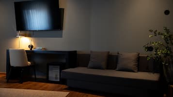 A modern living room with a sofa, a TV mounted on the wall, a desk with a chair, and a small plant.