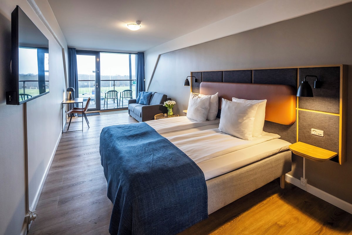 A modern hotel room with a large bed, a sofa, a TV, and a balcony with a view.