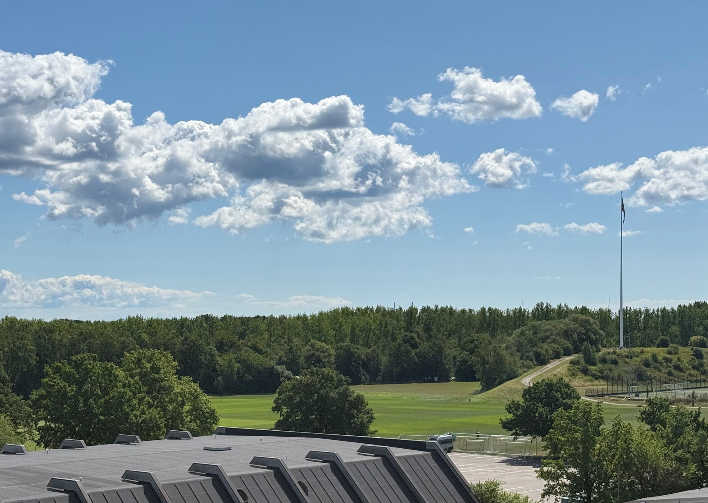 Cielo despejado con nubes dispersas, un campo verde y una colina boscosa al fondo.