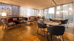 A spacious hotel lobby with a wooden floor, leather sofas, and modern chairs. There are large windows with curtains and pendant lights.