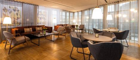 Un amplio vestíbulo de hotel con piso de madera, sofás de cuero y sillas modernas. Hay ventanas grandes con cortinas y lámparas colgantes.