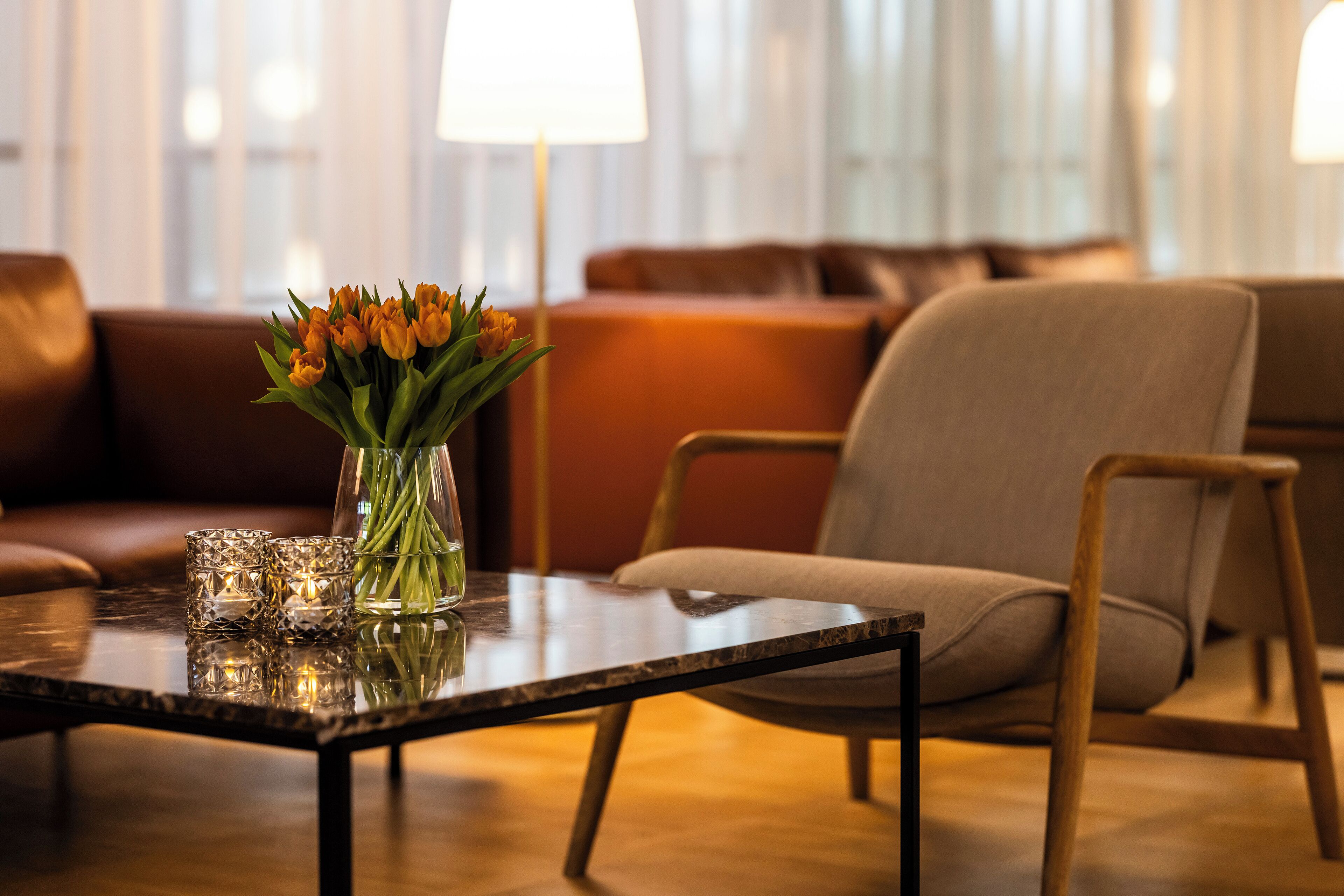 A modern living room with a glass vase of flowers on a glass table, a brown leather sofa, and a wooden chair.