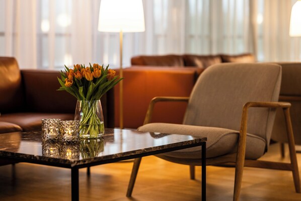 A modern living room with a glass vase of flowers on a glass table, a brown leather sofa, and a wooden chair. - Idraettens Hus Hotel & konference (Brondby)