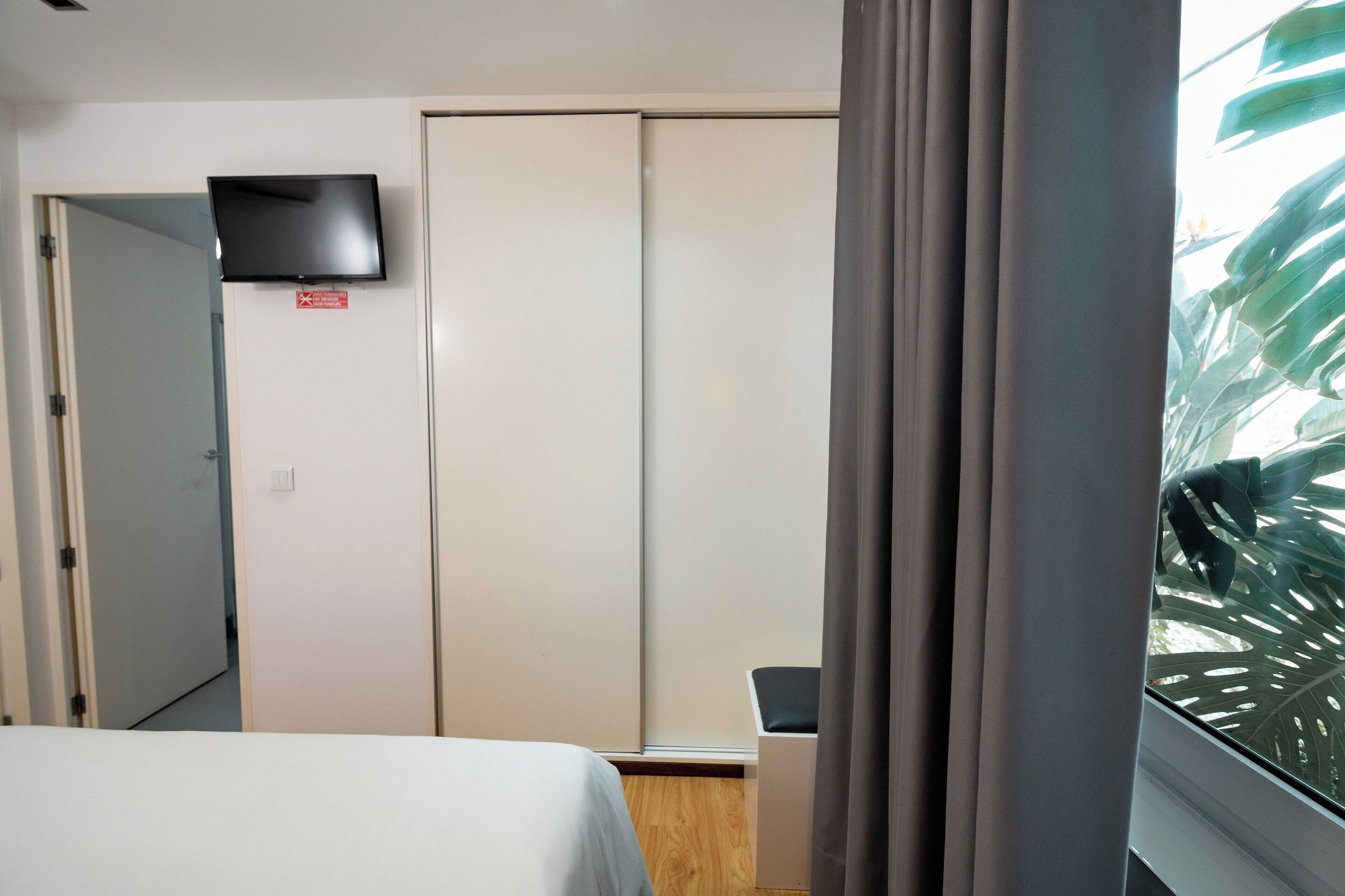 Habitación de hotel con cama, televisor montado en la pared, armario y ventana con cortinas.