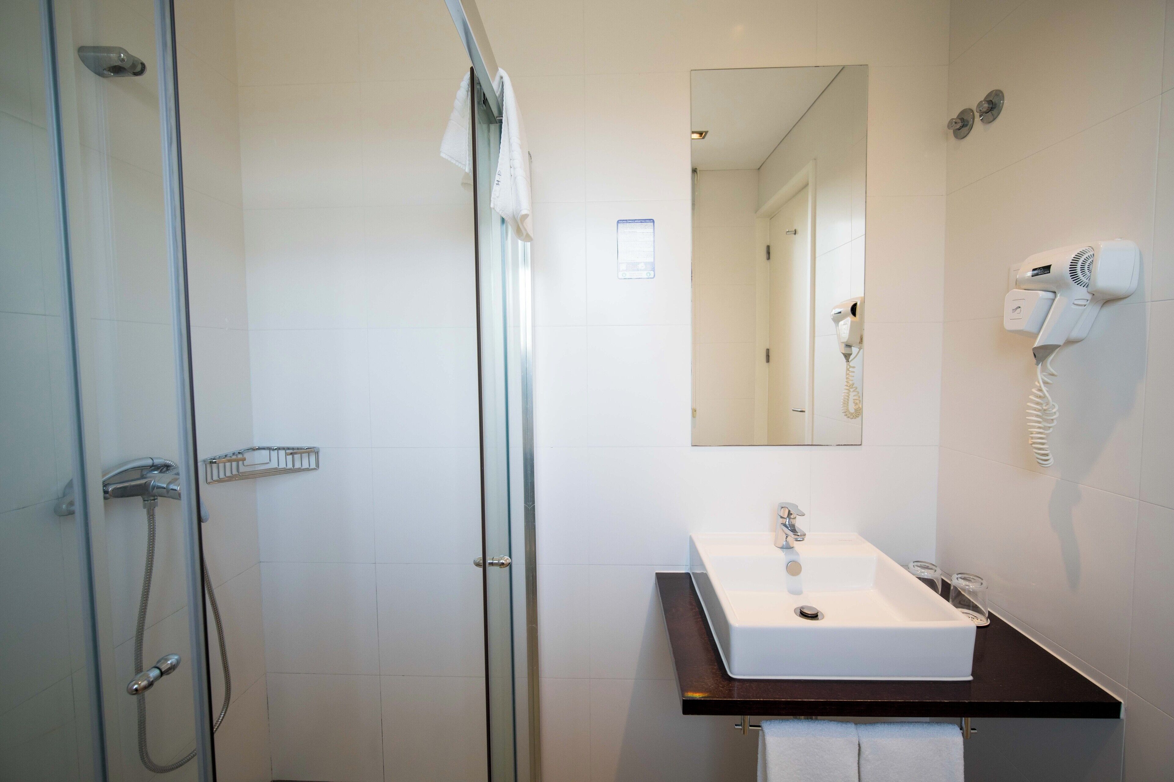 Un baño moderno con cabina de ducha de vidrio, un lavamanos blanco con encimera oscura y un secador de pelo montado en la pared.