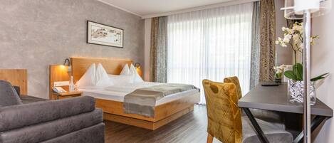 A modern hotel room with a wooden bed, a gray sofa, a dining table, and a large window with curtains.
