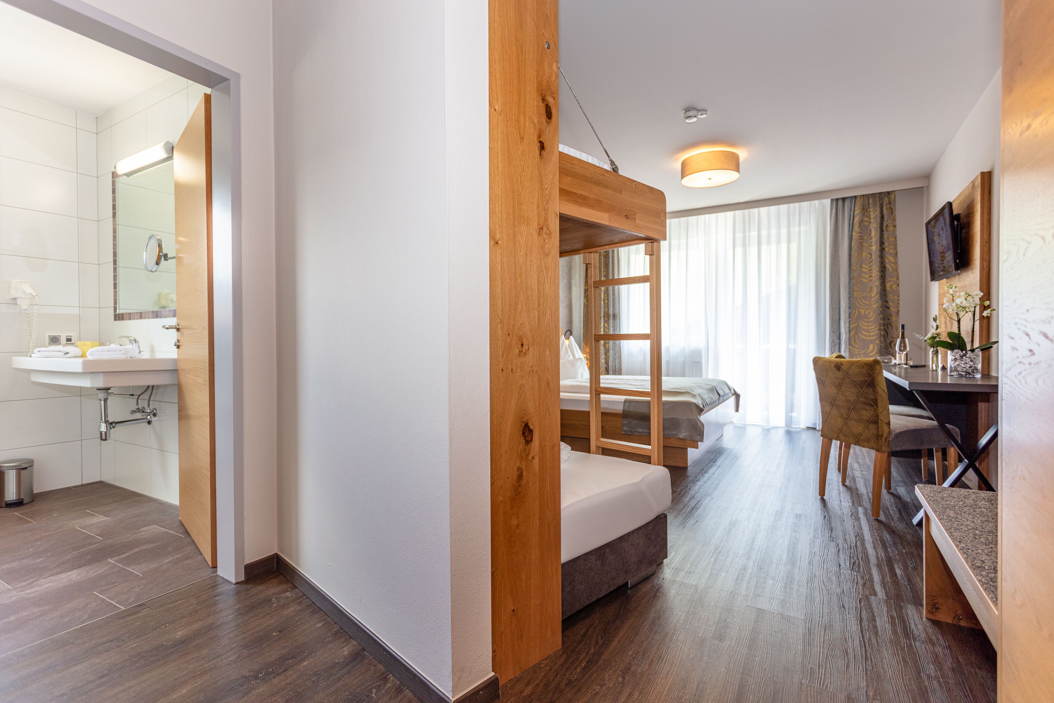 Una habitación de hotel con una cama nido de madera, un escritorio con silla, un lavabo y un espejo.