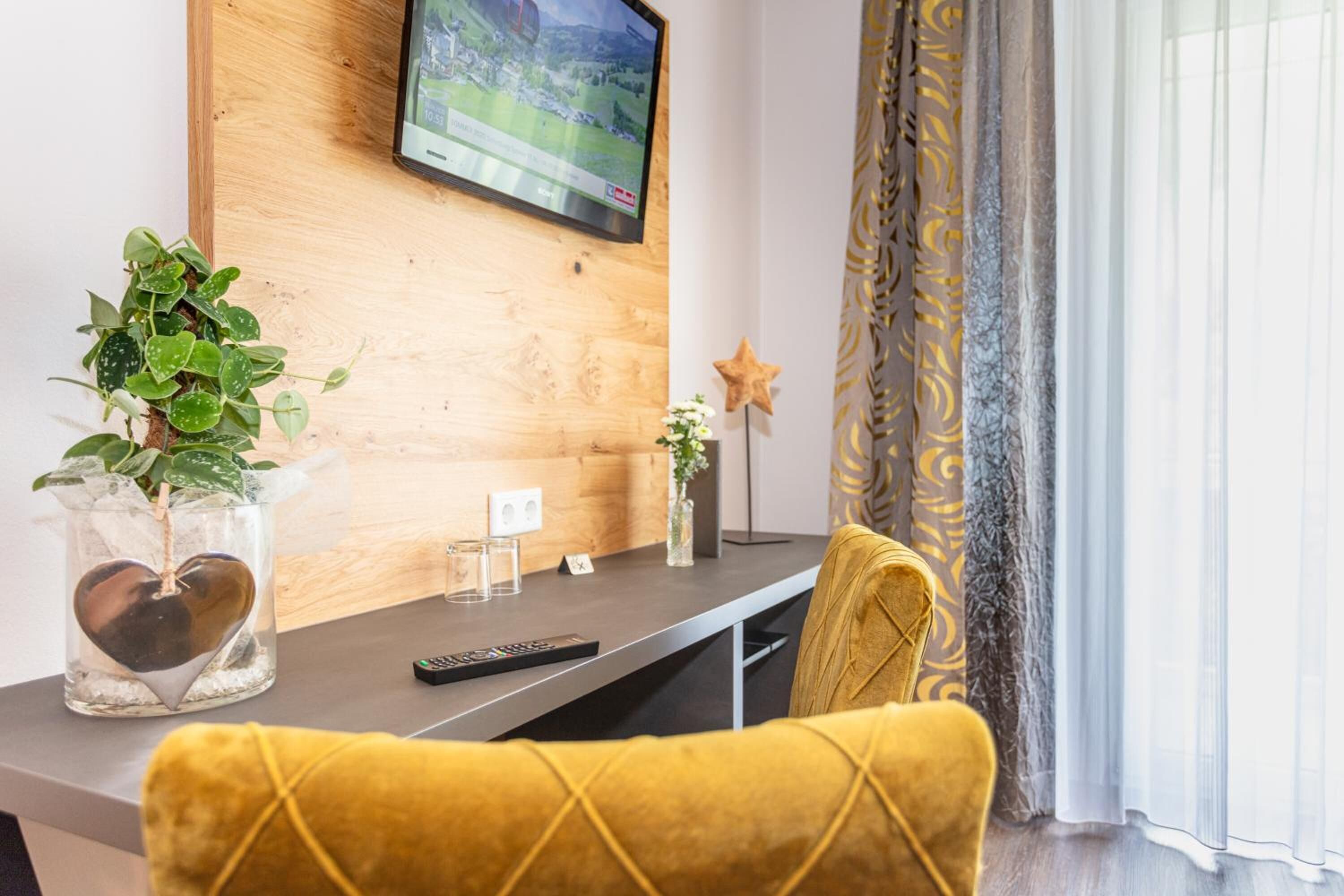 Un salon moderne avec un mur en bois, un téléviseur fixé au mur, un vase en verre orné d’un motif en forme de cœur, une plante en pot et un fauteuil jaune.